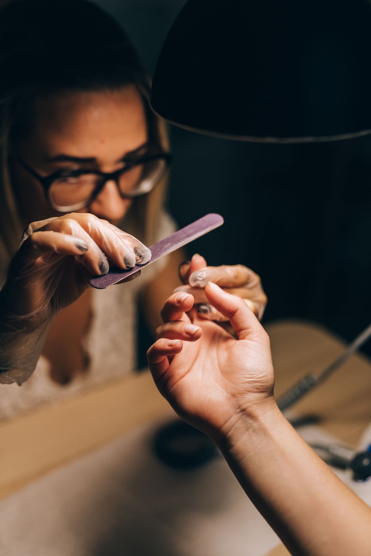 Nail technician carefully filing a client's nails under warm lamp light